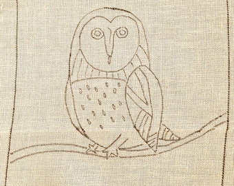 以下が含まれることがあります： ベージュ色のジュートキャンバスに描かれた、茶色のインクによるフクロウのイラスト。 フクロウは枝に止まっており、長方形の枠で囲まれています。 「twocatsanddoghooking」というテキストと「2026」という年号が下に書かれています。