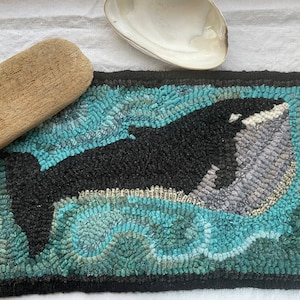 May include: Hand-hooked rug featuring a black and white orca whale swimming in turquoise waters. The rug has a black border and is rectangular. A piece of driftwood and a seashell are also in the image.