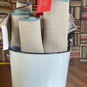 May include: A white enamel bucket with a black rim, filled with several rolls of beige linen fabric with a teal trim. The fabric is labeled "Two Cadence Hooking" and "Original Hand Hooked Rugs with a twist"
