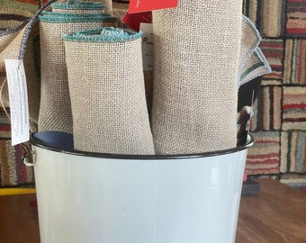 May include: A white enamel bucket with a black rim, filled with several rolls of beige linen fabric with a teal trim. The fabric is labeled "Two Cadence Hooking" and "Original Hand Hooked Rugs with a twist"