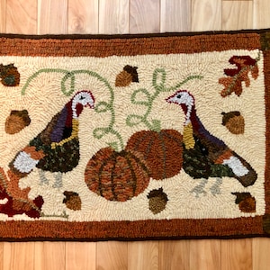 May include: A cream-colored hooked rug with a brown border features two turkeys, pumpkins, and acorns. The turkeys are brown, green, and purple with white accents. The pumpkins are orange and brown. The acorns are brown.