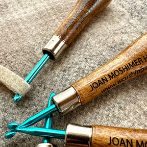 May include: Close-up of three rug hooking tools. Each tool has a wooden handle with the text "JOAN MOSHIMER HOOK" and a website address. The metal portion of the tools is a bright teal color. One tool is shown with a piece of fabric.