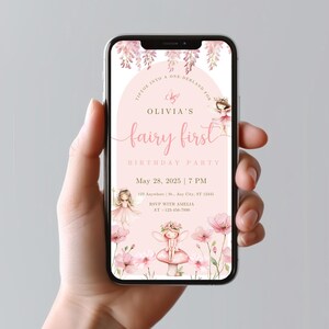 May include: A smartphone displays a pink invitation for Olivia's fairy-themed first birthday party. The invitation features floral and fairy illustrations, with the text "fairy first" in a cursive font. The date and time are also included.