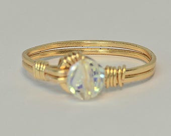 May include: A gold-colored wire-wrapped ring with a clear, faceted crystal bead at the center. The ring band is double-layered, and the crystal is secured with wire wrapping. The ring is set against a plain white background.