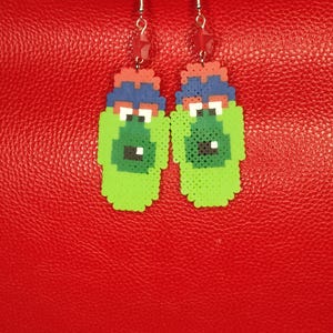 May include: A pair of handmade earrings featuring a pixelated green monster design. The earrings have a red and blue top, black and white eyes, and a black mouth. The earrings hang from silver hooks with a red bead.