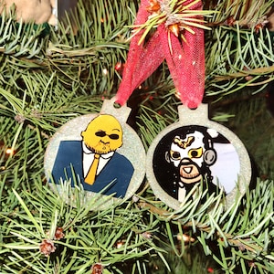 May include: Two round Christmas ornaments with glitter edges hang from a red ribbon on a Christmas tree. One ornament features a cartoon man in a suit and sunglasses. The other ornament has a black and white image of a man with a mask.