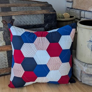 May include: A red, white and blue patchwork cushion with a hexagonal pattern. The cushion is made of different fabrics with various patterns, including polka dots and floral prints.