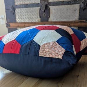 May include: A large, blue, red and white patchwork cushion with a navy blue border. The cushion is made of different fabrics and has a quilted design.
