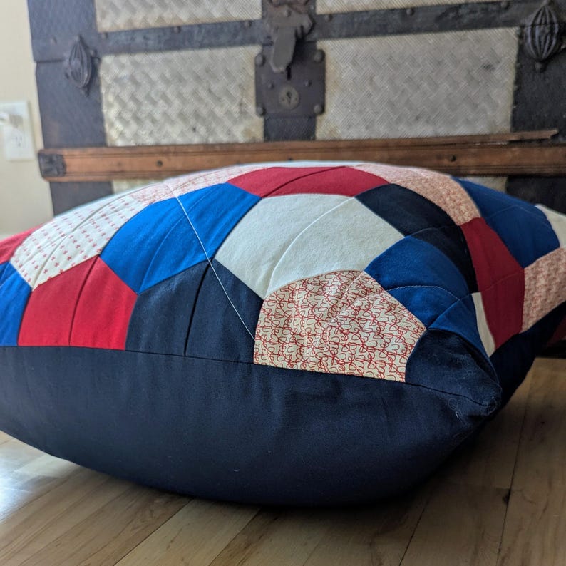 May include: A large, blue, red and white patchwork cushion with a navy blue border. The cushion is made of different fabrics and has a quilted design.