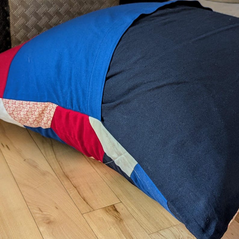 May include: A blue, red and white patchwork pillow with a dark blue fabric cover. The pillow is lying on a wooden floor.