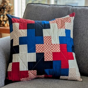 May include: A patchwork pillow with red, white, and blue squares. The pillow is made of different fabrics with various patterns, including gingham and polka dots.