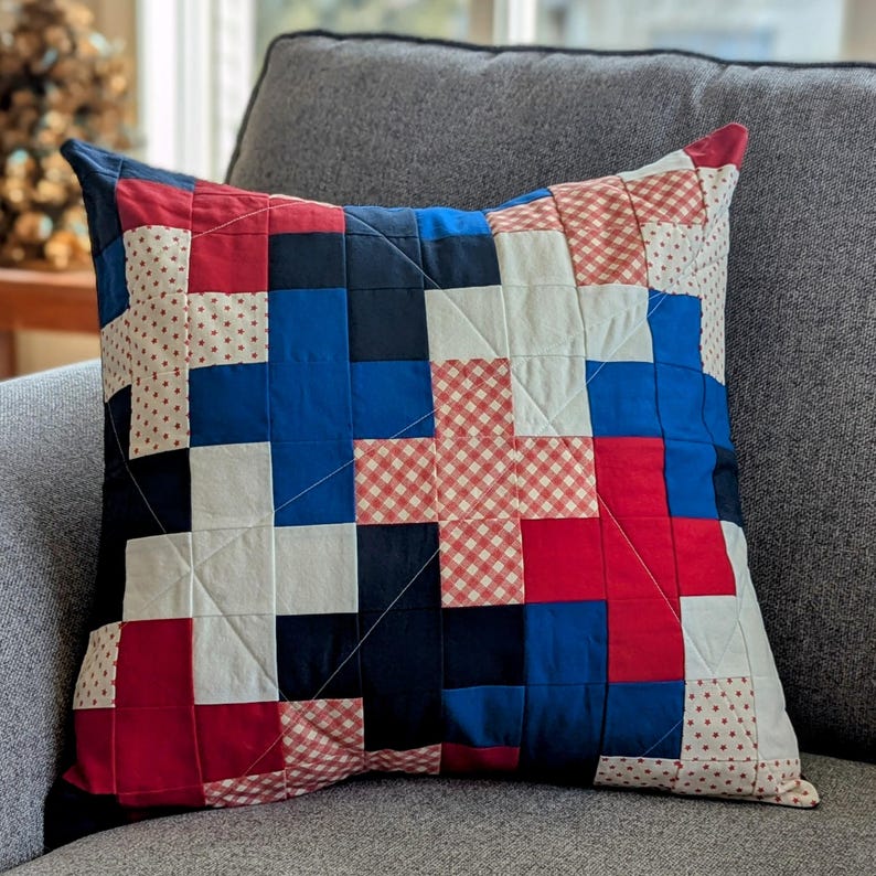 May include: A patchwork pillow with red, white, and blue squares. The pillow is made of different fabrics with various patterns, including gingham and polka dots.