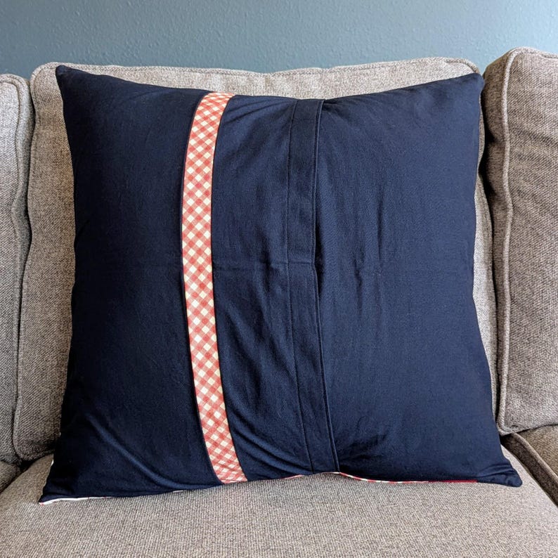 May include: A navy blue pillow with a red and white gingham trim. The pillow has a seam down the center and is made of a soft, comfortable fabric.