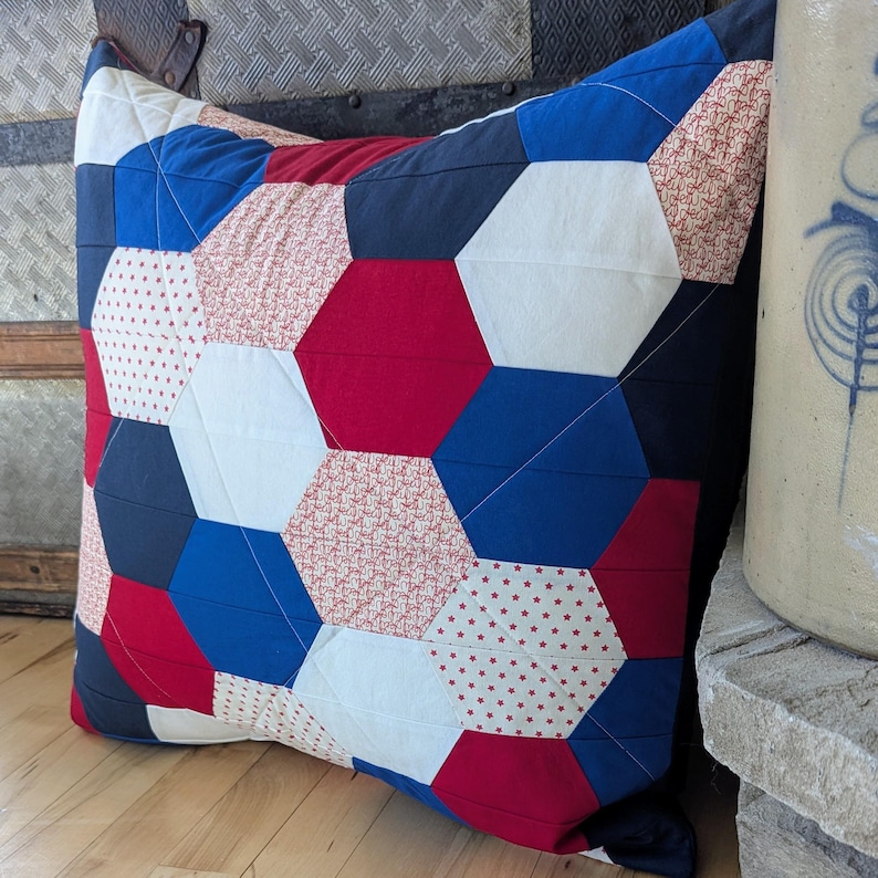 May include: A red, white and blue quilted cushion with a hexagonal pattern. The cushion is made of different fabrics with various patterns, including stars and polka dots.