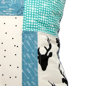 May include: A patchwork pillow cover with a white background and various patterns in blue, green, and black. The patterns include polka dots, arrows, and a silhouette of a deer head.