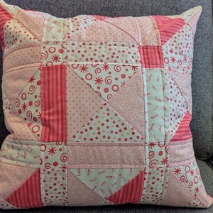 May include: Pink and white patchwork pillow with a geometric design. The pillow is made of different fabrics with various patterns, including polka dots, swirls, and stars. The fabric is stitched together to create a unique and eye-catching design. The pillow has the word "Love" written on it multiple times.