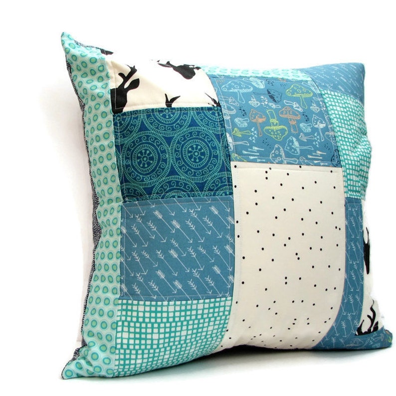 May include: A patchwork pillow with a blue and green color scheme. The pillow is made of different fabrics with various patterns, including circles, arrows, and polka dots. The pillow has a white background with black deer silhouettes.