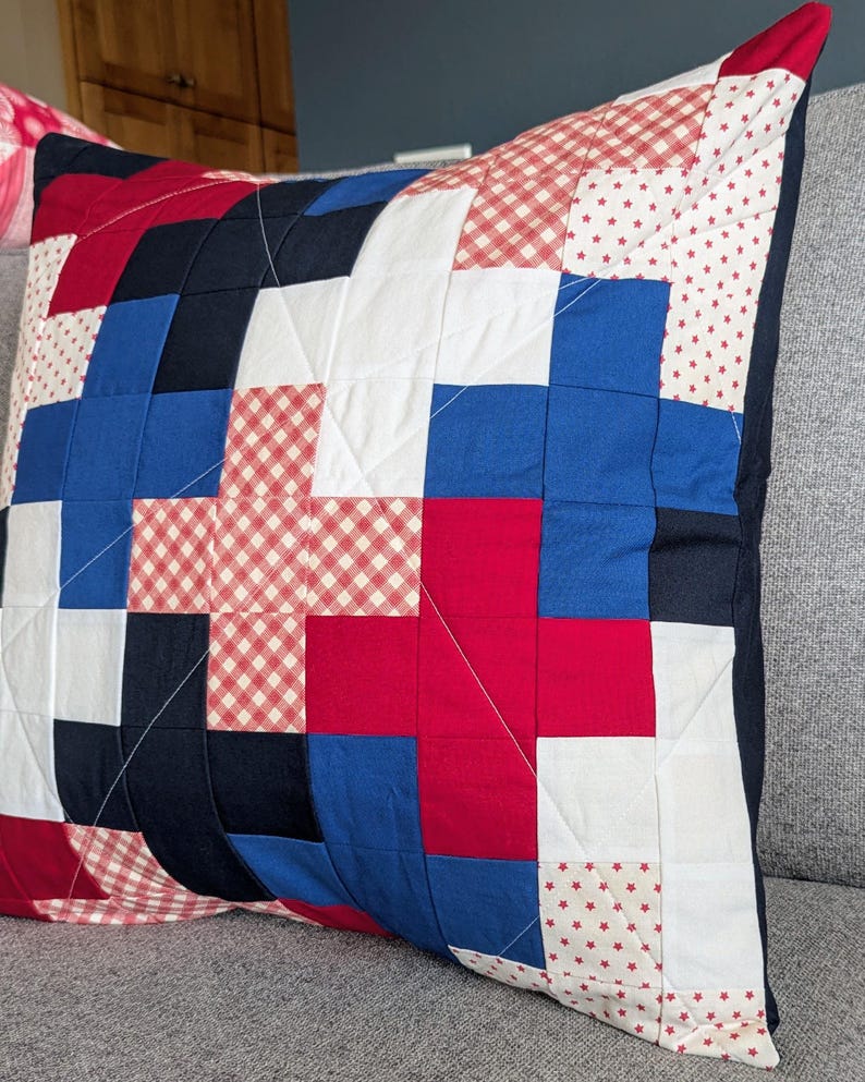 May include: A red, white, and blue patchwork pillow with a gingham pattern. The pillow is made of different fabrics and has a geometric design.