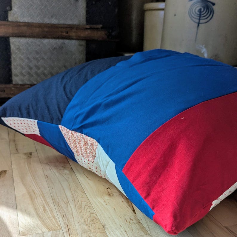 May include: A large, rectangular cushion with a patchwork design. The cushion is made of blue, red and white fabric with a geometric pattern. The cushion is lying on a wooden floor.