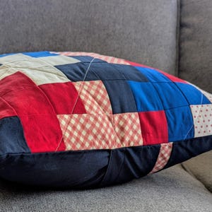 May include: A red, white, and blue patchwork pillow with a gingham pattern. The pillow is made of different fabrics and is quilted.
