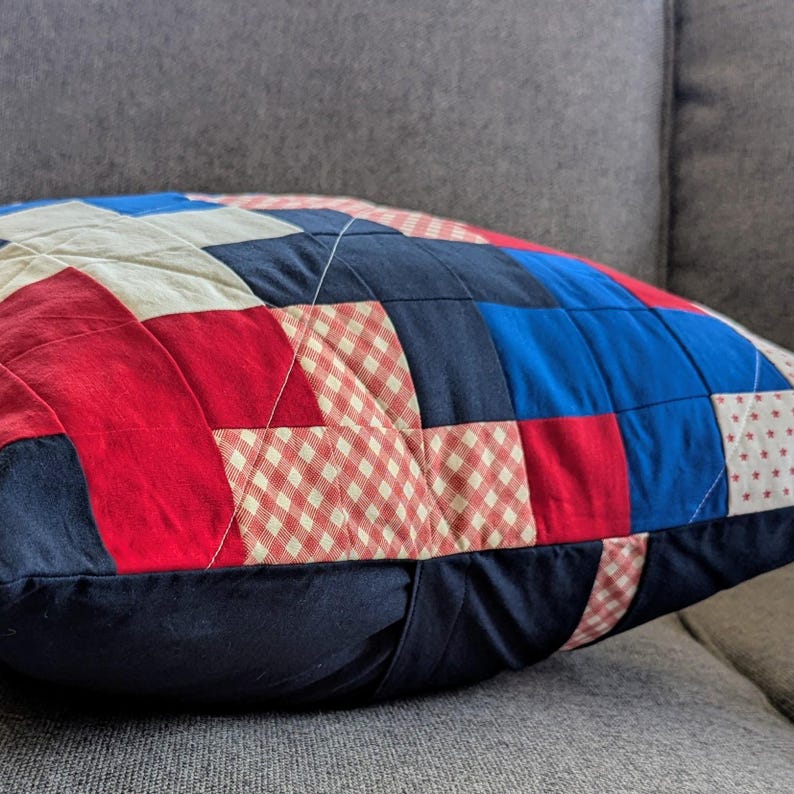 May include: A red, white, and blue patchwork pillow with a gingham pattern. The pillow is made of different fabrics and is quilted.
