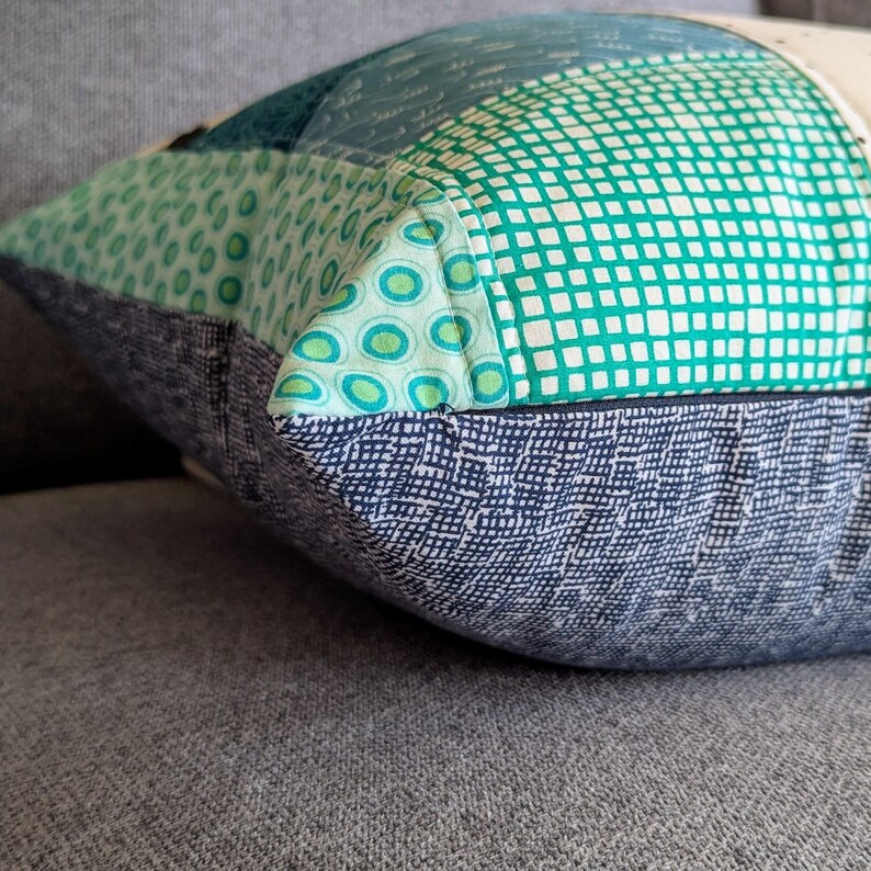 May include: Close-up of a pillow with a patchwork design featuring various green and blue fabrics. The pillow has a dark blue fabric with a subtle pattern on the back.