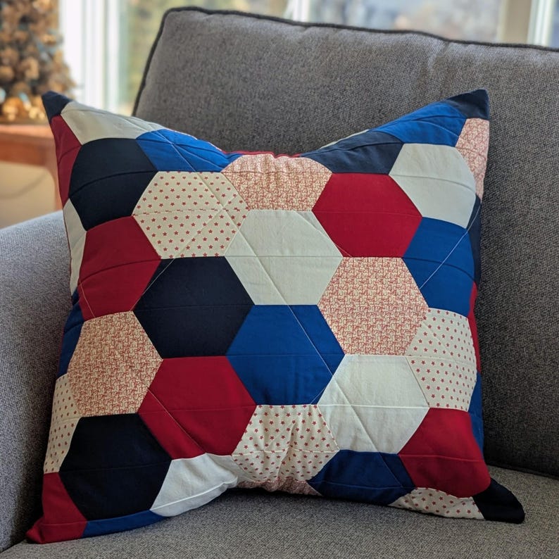 May include: A red, white and blue patchwork cushion with a hexagonal pattern. The cushion is made of different fabrics with various patterns, including polka dots and floral prints.