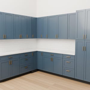 May include: An L-shaped kitchen cabinet set in a muted blue finish with shaker-style doors and drawers. The cabinetry features vertical brass-toned hardware pulls, a white countertop, and a white subway tile backsplash against a plain wall.