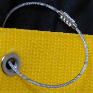 May include: Close-up of a silver metal cable loop with a textured clasp, attached to a yellow fabric with a metal grommet. The background is black. The cable is approximately 6 inches in diameter.