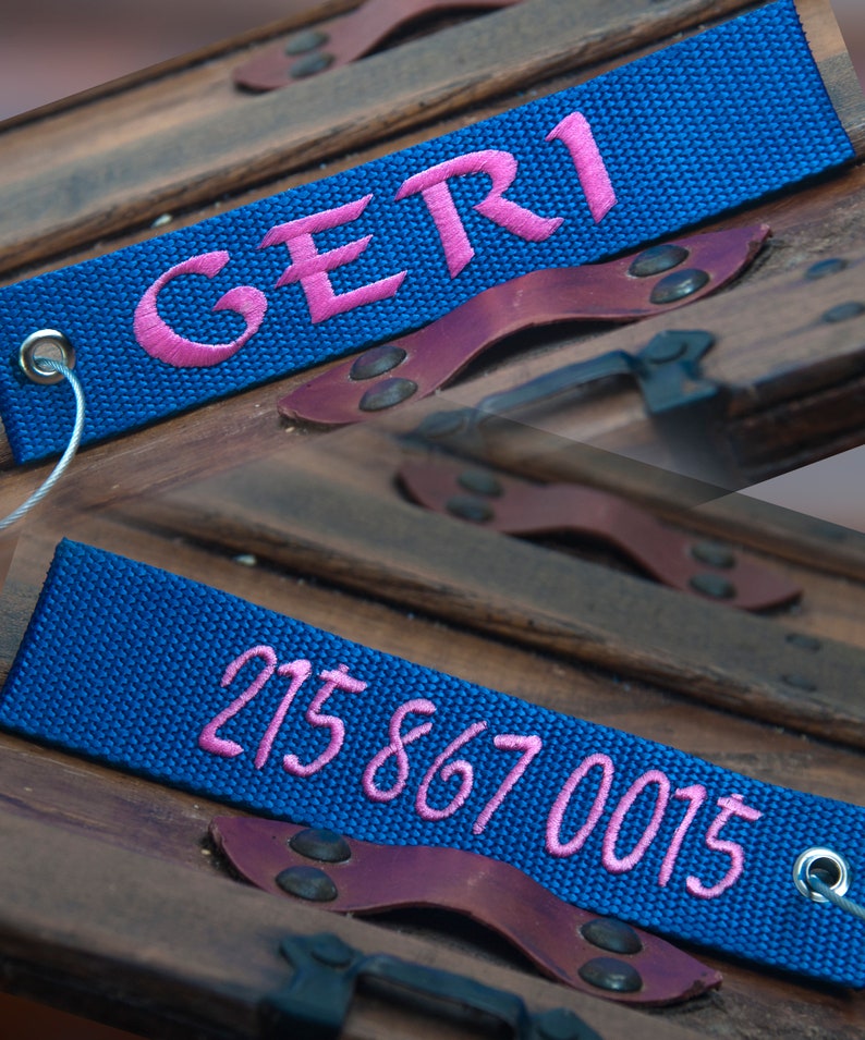 May include: Close-up of a blue luggage tag with the name "GERI" embroidered in pink. The tag is attached to a wooden surface with leather accents. Another tag displays the phone number "215 867 0015" in pink.