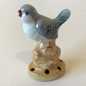 May include: A ceramic bird figurine with a light blue head and wings, a white chest, and a red beak. The bird is perched on a tan base with several holes. The figurine is likely a decorative item.