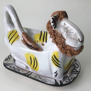 May include: A ceramic ram-shaped butter dish. The white dish features yellow and black stripes, a brown handle, and a decorative base. The ram has a brown mane and horns. A unique kitchen decor item.