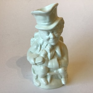 May include: White ceramic pitcher shaped like a person wearing a hat and sitting in a chair. The pitcher has a detailed face and is holding a small object in its hand.
