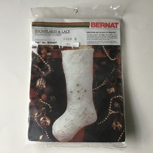 May include: A white stocking with a snowflake design, surrounded by gold and brown ornaments. The stocking is a counted stitchery pattern from Bernat, with the pattern number 1456 H and the kit number W09297.