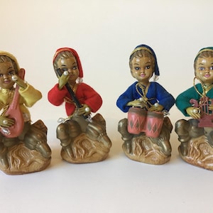 May include: Four ceramic figurines of musicians sitting on textured bases. Each figure wears a colored felt hat and matching long-sleeved top in yellow, red, blue, or green. They hold miniature instruments including a lute, flute, drums, and lyre.