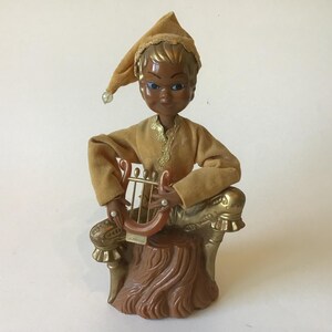 May include: A vintage figurine of a seated figure playing a lyre. The figure has dark skin, blue eyes, and wears a gold-colored outfit with a matching hat. The figurine is seated on a brown tree stump base.