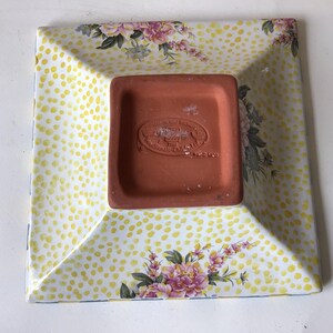 May include: Square ceramic dish with a yellow polka dot pattern and floral accents. The dish has a terracotta-colored base with embossed text. The edges are slightly raised, and the corners are angled.
