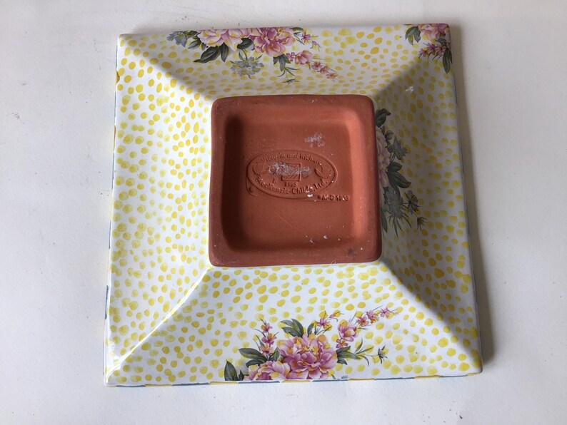 May include: Square ceramic dish with a yellow polka dot pattern and floral accents. The dish has a terracotta-colored base with embossed text. The edges are slightly raised, and the corners are angled.