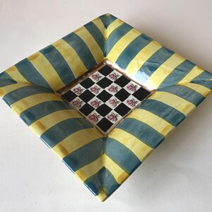 May include: Square decorative ceramic dish with a unique design. The dish features alternating yellow and teal blue stripes on the outer edges. The center of the dish has a black and white checkered pattern with pink floral accents.