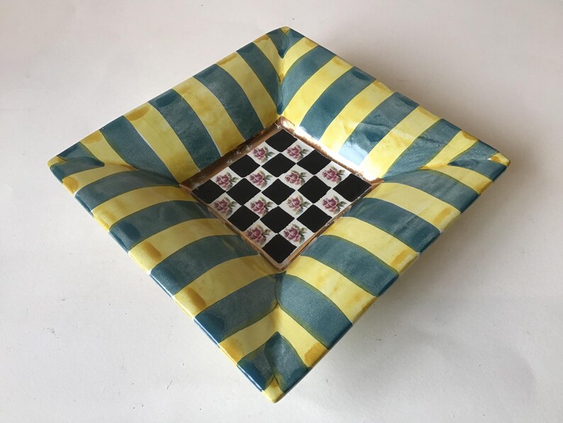 May include: Square decorative ceramic dish with a unique design. The dish features alternating yellow and teal blue stripes on the outer edges. The center of the dish has a black and white checkered pattern with pink floral accents.