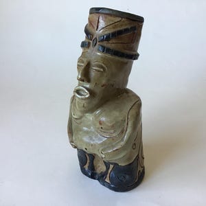 May include: A handcrafted ceramic bust of a figure with a stylized face and a detailed headdress. The sculpture features a muted color palette of tan, brown, and black, with a smooth finish. The figure's expression is serene, and the overall aesthetic is reminiscent of ancient art.