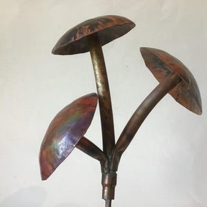 May include: A handcrafted metal sculpture featuring three stylized mushroom forms. The caps have a reddish-brown patina with hints of green and purple. The stems are a darker bronze color, creating a naturalistic garden accent.