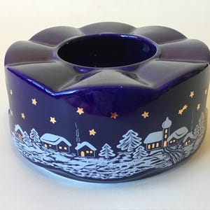 May include: A dark blue ceramic candle holder with a winter village scene. The holder has a scalloped top and is decorated with gold stars and a white and blue village design around the base.