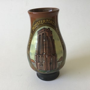 May include: A brown ceramic vase with a painted image of a church tower. The vase has the words "MONSTERTOREM" and "ST. LIEVENS" written on it.