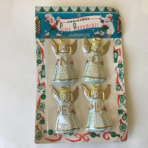 May include: A set of four vintage-style ceramic angel ornaments packaged on a cardboard backing. Each angel features blonde hair, gold wings, and a white dress with a gold star pattern. The angels hold the letters N, O, E, and L in gold.
