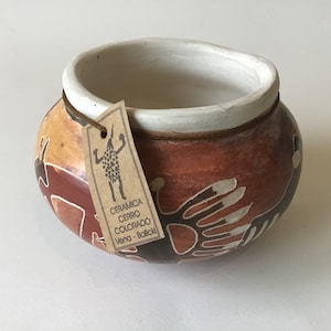 May include: Hand-painted ceramic bowl with a brown, orange, and white color scheme. The bowl features a unique design with a tag attached. The tag has text and a drawing of a person. The bowl is a decorative item.
