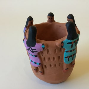 May include: Terracotta planter with five figures painted in blue, pink, and black, holding hands around the rim of the planter.
