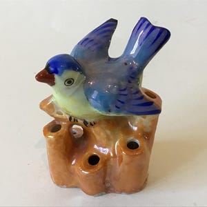 May include: A ceramic bird figurine with a blue head and wings, perched on a brown tree stump with three holes. The bird has a yellow chest and a red beak. A decorative item.