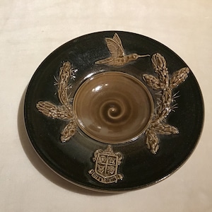 May include: A dark green ceramic plate with a central brown bowl featuring a spiral design. The plate is decorated with a hummingbird, cactus-like designs, and a crest with the words "DEDITA DEUS".