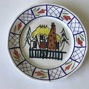 May include: A white ceramic plate with a blue and white checkered border. The center of the plate features a hand-painted illustration of a brown and yellow cottage with a thatched roof, a chimney, and a fence. The cottage is surrounded by green trees.